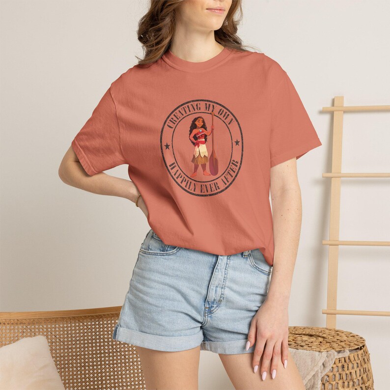 Op de afbeelding: Koraalrood T-shirt met een cirkelvormige afbeelding met de tekst "Creating My Own Happily Ever After". De afbeelding bevat een illustratie van een personage met een peddel. Het shirt wordt gecombineerd met een spijkerbroek.