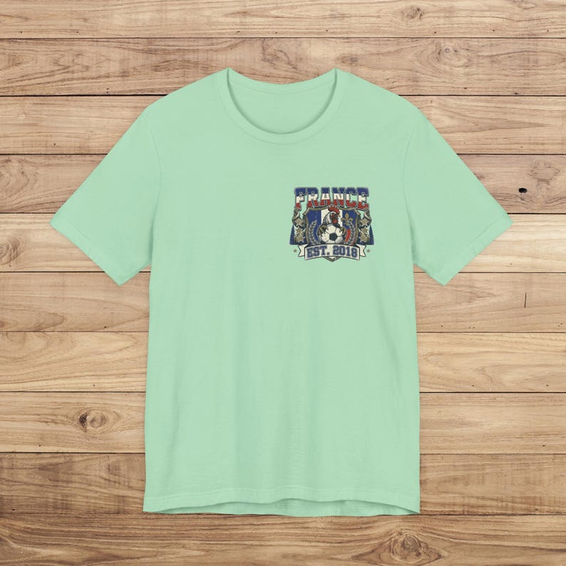 May include: A light mint green short-sleeve t-shirt featuring a small graphic on the left chest. The design includes a rooster, a soccer ball, and the text "FRANCE" above and "EST. 2018" below, all rendered in red, white, and blue colors.