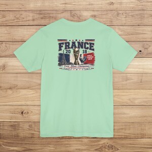 May include: A light mint green t-shirt featuring a graphic on the back with the text FRANCE 2018, WORLD CHAMPIONS 2018, and FIERS, BLEUS, CHAMPIONS. The design includes the Eiffel Tower, the World Cup trophy, a rooster, and the French flag.