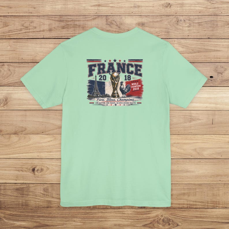 May include: A light mint green t-shirt featuring a graphic on the back with the text FRANCE 2018, WORLD CHAMPIONS 2018, and FIERS, BLEUS, CHAMPIONS. The design includes the Eiffel Tower, the World Cup trophy, a rooster, and the French flag.