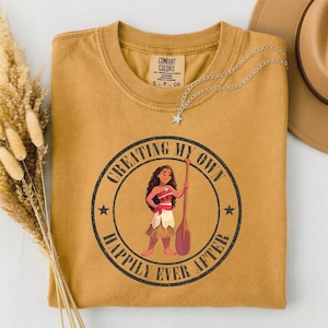 Op de afbeelding: Mosterdkleurig T-shirt met een afbeelding van een vrouw met een peddel, omringd door de tekst "CREATING MY OWN HAPPILY EVER AFTER". Een zilveren sterrenketting en een beige hoed zijn ook zichtbaar.
