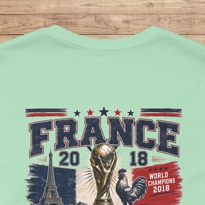 May include: A light green t-shirt featuring a graphic print with the text FRANCE 2018, a depiction of the Eiffel Tower, a soccer trophy, a rooster, and the text WORLD CHAMPIONS 2018, accented with red, white, and blue stars and stripes.