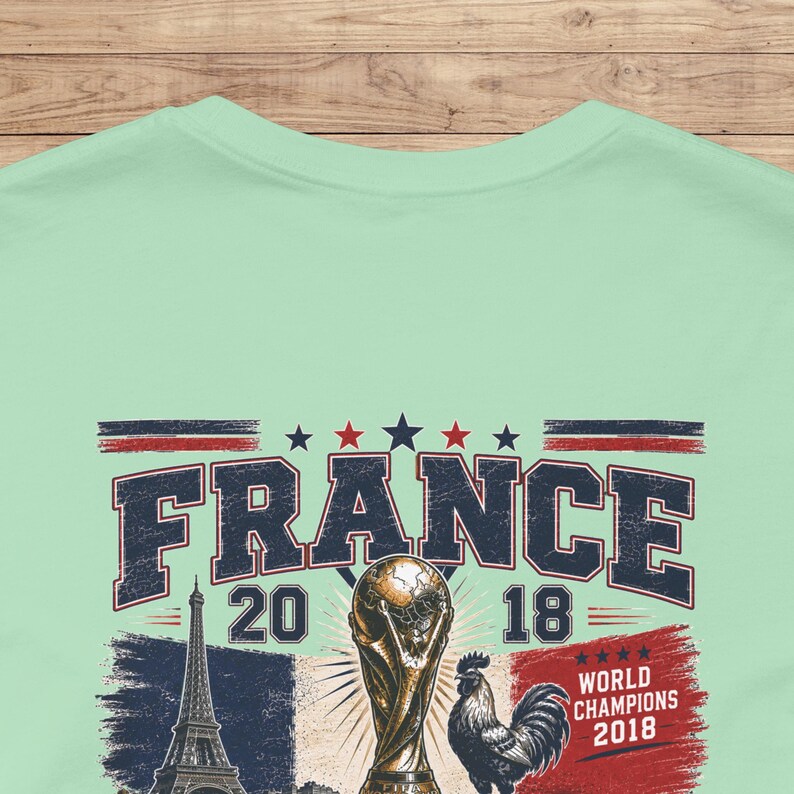 May include: A light green t-shirt featuring a graphic print with the text FRANCE 2018, a depiction of the Eiffel Tower, a soccer trophy, a rooster, and the text WORLD CHAMPIONS 2018, accented with red, white, and blue stars and stripes.