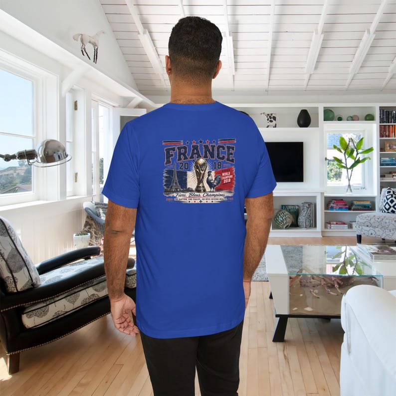 May include: A royal blue short-sleeve t-shirt featuring a graphic on the back. The design includes the text FRANCE 2018, an illustration of the Eiffel Tower, the World Cup trophy, a rooster, and the text Fiers, Bleus, Champions.