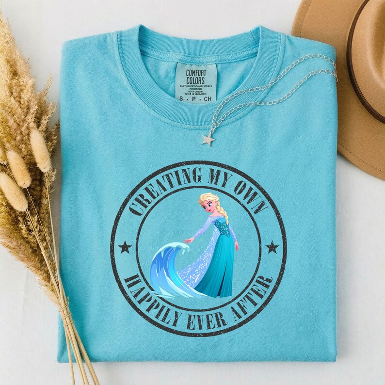Puede incluir: Una camiseta azul claro con un gr&aacute;fico redondo que presenta una princesa de dibujos animados y el texto "CREATING MY OWN HAPPILY EVER AFTER". Un collar plateado con un colgante de estrella est&aacute; sobre la camiseta.