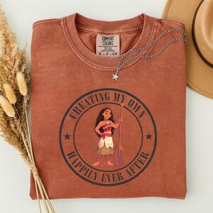 Op de afbeelding: Een roestkleurig t-shirt met een afbeelding van een stripfiguur met een peddel, met de tekst "CREATING MY OWN HAPPILY EVER AFTER" in een cirkelvormig ontwerp. Een zilveren ketting met een sterhanger en een bruine hoed zijn ook zichtbaar.