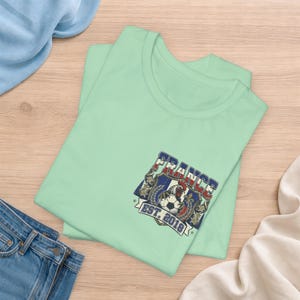 May include: A light green t-shirt featuring a graphic on the chest. The design includes a shield with a rooster, a soccer ball, and the text FRANCE EST. 2018 in blue, white, and red colors. The graphic is framed by decorative elements and stars.