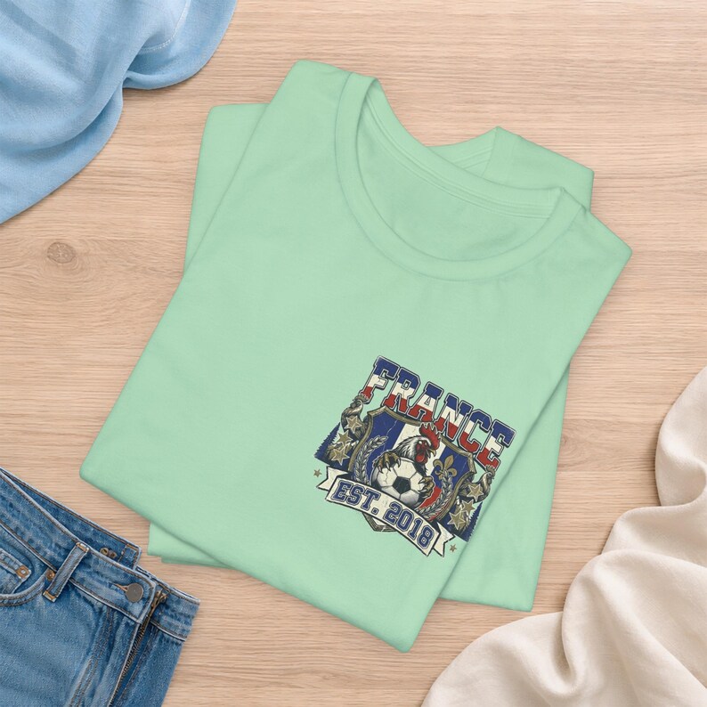 May include: A light green t-shirt featuring a graphic on the chest. The design includes a shield with a rooster, a soccer ball, and the text FRANCE EST. 2018 in blue, white, and red colors. The graphic is framed by decorative elements and stars.
