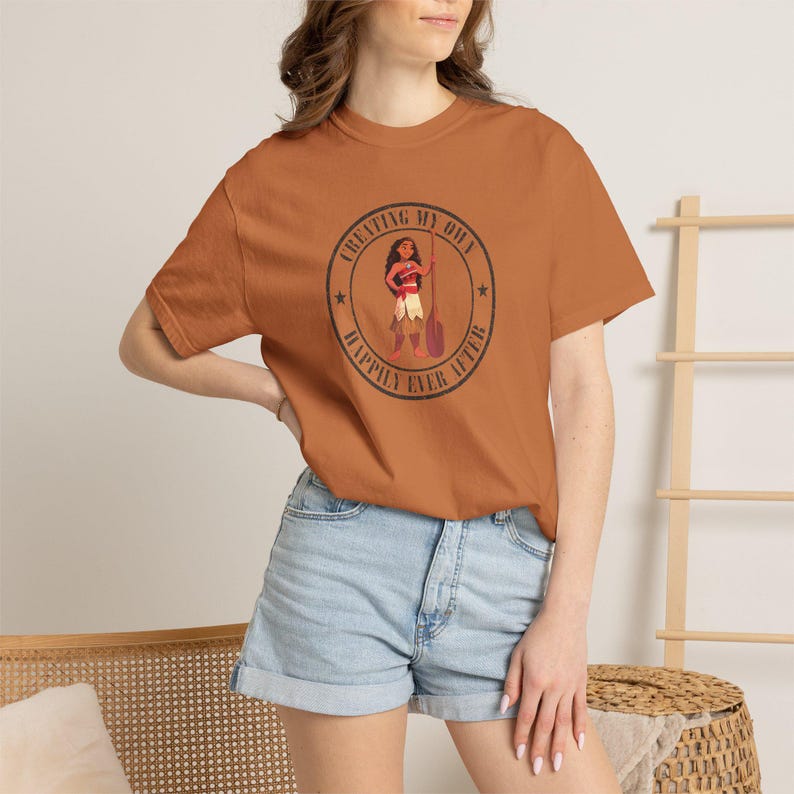 Op de afbeelding: Een roestkleurig T-shirt met een cirkelvormige afbeelding van een stripfiguur met een peddel. De afbeelding bevat de tekst "CREATING MY OWN HAPPILY EVER AFTER". Het shirt wordt gecombineerd met een spijkerbroekshort.