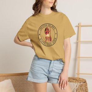 Op de afbeelding: Mosterdgeel t-shirt met een cirkelvormige afbeelding met een stripfiguur met een peddel. De afbeelding bevat de tekst "CREATING MY OWN" boven het personage en "HAPPILY EVER AFTER" eronder.