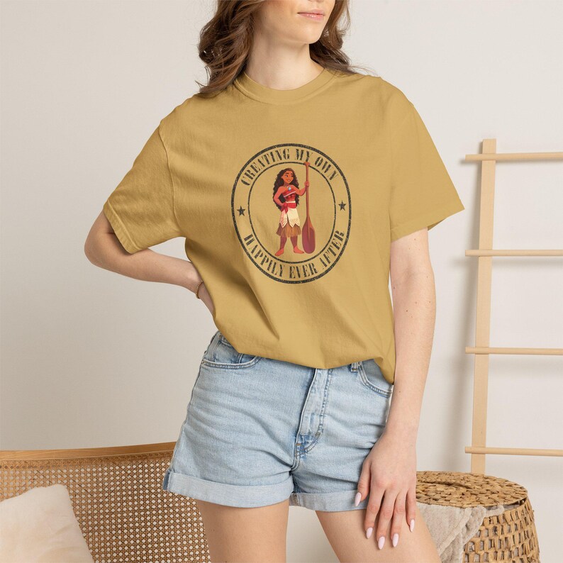 Op de afbeelding: Mosterdgeel t-shirt met een cirkelvormige afbeelding met een stripfiguur met een peddel. De afbeelding bevat de tekst "CREATING MY OWN" boven het personage en "HAPPILY EVER AFTER" eronder.