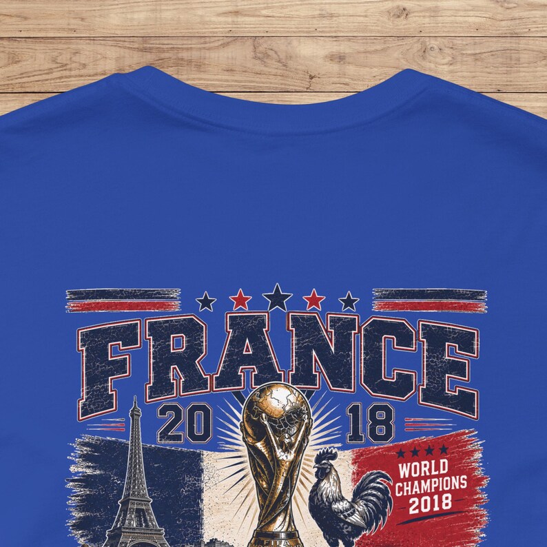 May include: A royal blue t-shirt featuring a graphic print with the text FRANCE 2018. The design includes an illustration of the Eiffel Tower, a gold-colored trophy, a rooster, and the text WORLD CHAMPIONS 2018 with four stars above the text.