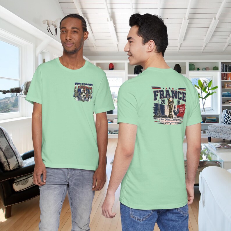 May include: A light green t-shirt featuring a small graphic on the front chest and a large graphic on the back. The back design displays the text FRANCE 2018, an illustration of the Eiffel Tower, a trophy, and a soccer player with the text World Champions.