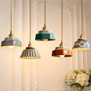 May include: Five pendant lights with ceramic shades in various colours and patterns, including blue, green, and orange. Each light has a brass fixture and a decorative cord, creating a warm ambiance. The lights are hanging in front of a white wall.