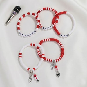 May include: A collection of red and white beaded bracelets with charms and lettering spelling out "ARIRANG", "ARMY", and "BTS". A silver microphone is in the upper left corner. The bracelets have various charms, including a heart, a hand gesture, and a cross.