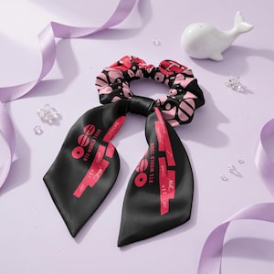 Pode incluir: Uma scrunchie preta com um padrão floral rosa e preto e caudas pretas longas. As caudas têm o texto "BTS WORLD TOUR" em vermelho. Uma estatueta de baleia branca e fita roxa estão no fundo.