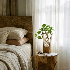 Pode incluir: Um vaso de madeira com uma inserção de vidro que contém uma planta verde com folhas em forma de coração. O vaso está sobre uma mesa lateral rústica de madeira ao lado de uma cama com roupa de cama de cores neutras. Cortinas brancas transparentes emolduram a janela.
