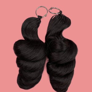 Peut inclure: Deux extensions de cheveux noirs avec des anneaux argentés, sur fond rose. Les extensions sont coiffées en boucles ondulées volumineuses, d'une couleur sombre et riche. Les anneaux sont fixés en haut des extensions.