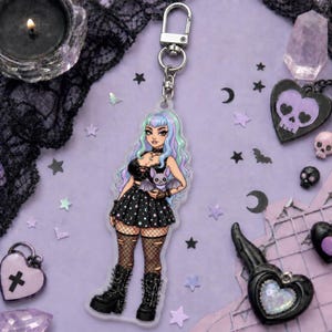 May include: A clear acrylic keychain featuring a gothic anime girl with pastel hair, holding a bat. She wears a black dress with moon and star details, fishnet stockings, and platform boots. The keychain has a silver clasp.