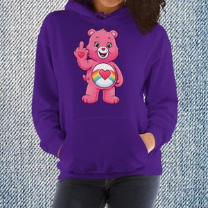 May include: Purple hoodie featuring a pink cartoon bear with a rainbow heart on its belly and giving the middle finger. The bear has a heart on its paw. The hoodie has a kangaroo pocket and a drawstring hood.