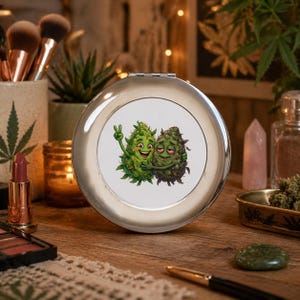 May include: A silver compact mirror with a cartoon illustration of two cannabis buds hugging. One bud is green and giving a peace sign, while the other is dark green. The mirror is on a wooden surface with makeup and accessories.