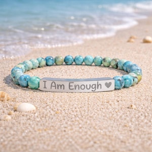May include: A beaded bracelet with turquoise and blue spherical beads. A silver bar with the inscription "I Am Enough" and a heart symbol is centered on the bracelet. The bracelet is resting on a sandy beach with the ocean in the background.