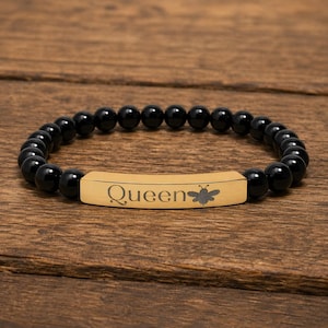 May include: Black beaded bracelet with a gold-colored rectangular centerpiece. The centerpiece is engraved with the word "Queen" and a bee graphic. The bracelet is displayed on a wooden surface.