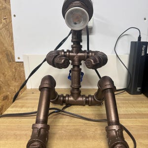 May include: A handmade industrial-style lamp constructed from copper-colored metal pipes and fittings. The lamp features a round light fixture at the top and is designed to resemble a person sitting. The lamp is plugged into an electrical outlet.
