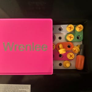 May include: A bright pink square storage box with the name Wrenlee printed in green on the lid. The box is open, revealing a white pegboard base inside containing various colorful plastic construction pieces, including orange, green, and purple bars, yellow screws, and an orange screwdriver tool.