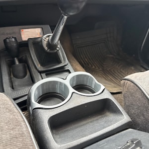 May include: Interior view of a vehicle's console, featuring a manual gear shift with a black knob and a silver shaft. A gray cup holder with two circular openings is positioned in front of the gear shift. The console is black and gray.