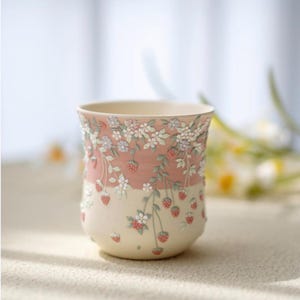 May include: A ceramic mug with a cream interior and a two-tone exterior. The top half is a soft pink, and the bottom half is cream. The mug is decorated with a raised floral and strawberry design, featuring white flowers and red strawberries.