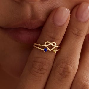 May include: A gold ring featuring a central heart-shaped wire design. The band splits into two thin lines, each accented with a small round gemstone. One stone is a deep blue, and the other is a bright yellow, set symmetrically on the band.