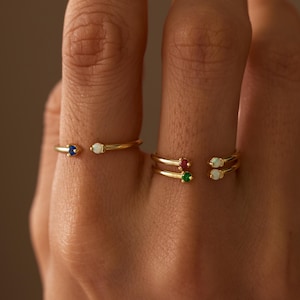 May include: Two gold open-style rings worn on fingers. The first ring features a small blue gemstone and a white opal. The second ring features a red gemstone, a green gemstone, and two white opals. The text CaeloriJewelryCo is in the corner.