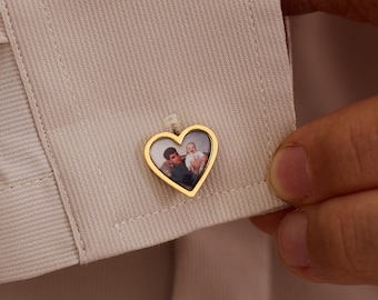 May include: Gold heart-shaped cufflink with a photograph inside, attached to a light beige dress shirt. The cufflink is being held by a person's hand. The photograph appears to be of two people. The cufflink is a personalized jewelry item.