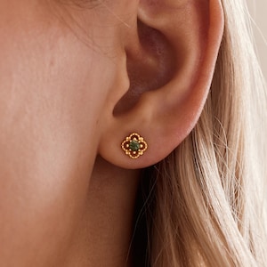 May include: A gold stud earring featuring a four-lobed, clover-like frame with a beaded edge and a small, round, dark green gemstone set in the center. The earring is positioned on an earlobe, showcasing its intricate metalwork and gemstone.