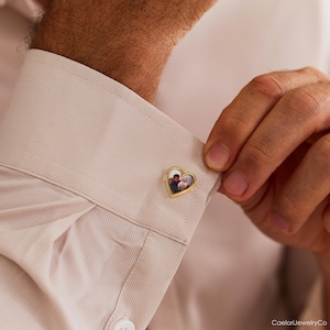 May include: Gold-toned heart-shaped cufflink with a personalized photo. The cufflink is attached to a light beige dress shirt. The photo inside the heart shows two people.