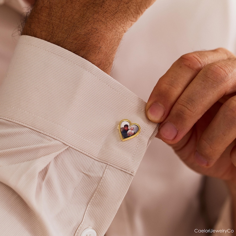 May include: Gold-toned heart-shaped cufflink with a personalized photo. The cufflink is attached to a light beige dress shirt. The photo inside the heart shows two people.