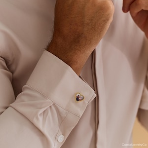 May include: Gold-toned heart-shaped cufflink with a small photo inside. The cufflink is on a light beige dress shirt. The photo inside the heart appears to be of two people. The cufflink is a personalized jewelry item.
