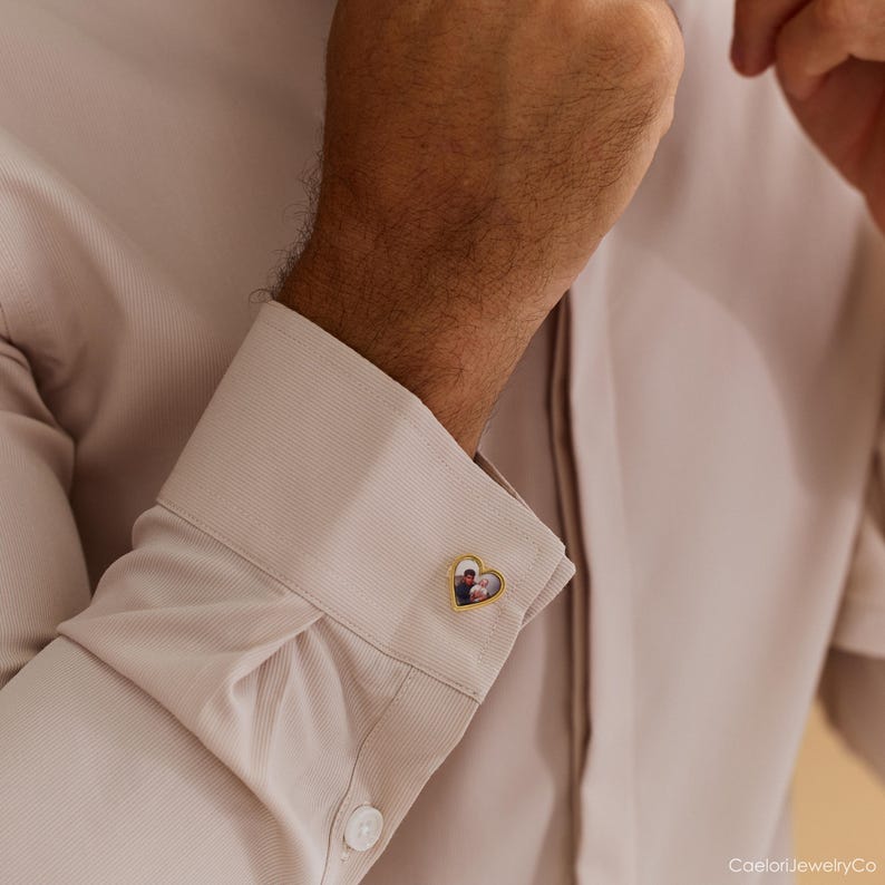 May include: Gold-toned heart-shaped cufflink with a small photo inside. The cufflink is on a light beige dress shirt. The photo inside the heart appears to be of two people. The cufflink is a personalized jewelry item.