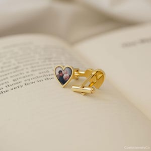 May include: Gold-colored cufflinks with a heart-shaped design. Each cufflink features a small, heart-shaped photo. The cufflinks are resting on the open pages of a book. The cufflinks are a classic accessory.