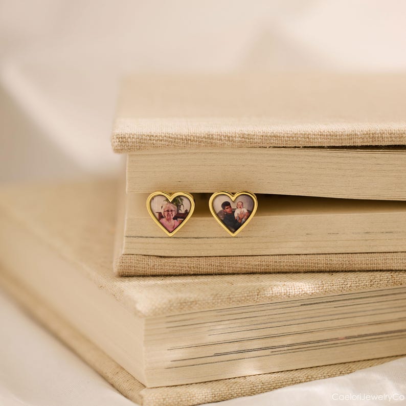 May include: Two gold heart-shaped earrings with personalized photos. The earrings are displayed on a stack of beige books. The earrings have a gold border and a photo inside. The earrings are a personalized jewelry item.
