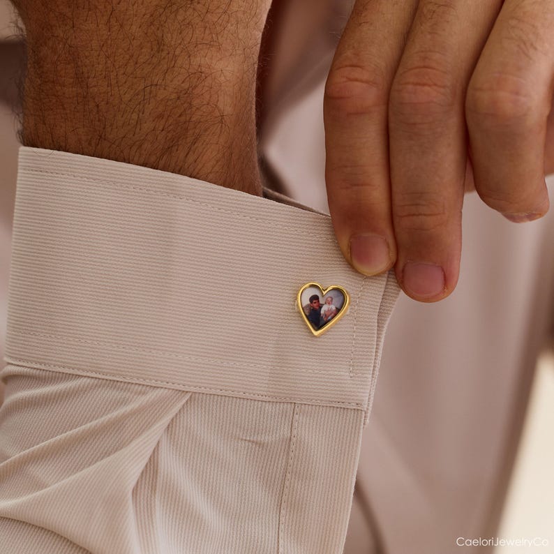 May include: Gold-colored heart-shaped cufflink with a photograph inside. The cufflink is on a light beige dress shirt cuff. A hand is adjusting the cufflink. The photograph inside the heart appears to be of a couple.