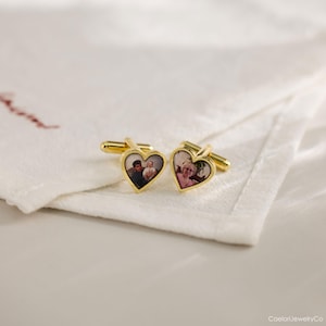 May include: A pair of gold-toned cufflinks, each featuring a heart-shaped design with a photograph inside. The cufflinks are displayed on a white cloth. The heart-shaped design is filled with a photograph of a person.