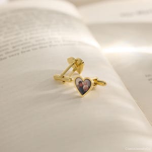 May include: Gold-colored cufflinks with a heart-shaped design. Each cufflink features a small, framed photograph of two people. The cufflinks are resting on an open book, suggesting a sentimental or personalized gift.