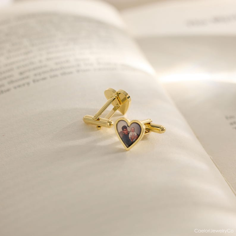 May include: Gold-colored cufflinks with a heart-shaped design. Each cufflink features a small, framed photograph of two people. The cufflinks are resting on an open book, suggesting a sentimental or personalized gift.