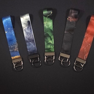 May include: Five keychains with different designs. The keychains have a metal clasp and a fabric strap. The straps feature designs of a starry night, clouds, green marble, black marble, and red marble.