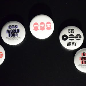 May include: A collection of white round buttons with various designs and text. Some buttons feature the text "BTS WORLD TOUR" and "ARLINGTON.TX 2026.08.15-16". Other buttons have the text "BTS ARMY" and a graphic.