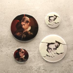 May include: Four circular pin-back buttons featuring photographic imagery. Two buttons display a person with light hair in a dark setting, while the other two buttons show a black and white close-up of eyes and hands peering through a torn paper effect.