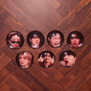Puede incluir: Un conjunto de siete chapas redondas, cada una con un retrato fotográfico distinto de una persona diferente con diversas expresiones faciales, incluyendo una persona con gafas de sol oscuras y otra haciendo un gesto de doble pulgar hacia arriba.