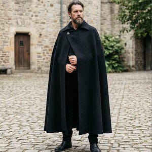 May include: A floor-length black wool cape featuring a structured collar and a single metallic button closure at the neck. The garment is worn over a black shirt and black trousers, with the fabric draping down to the ankles in a full silhouette.